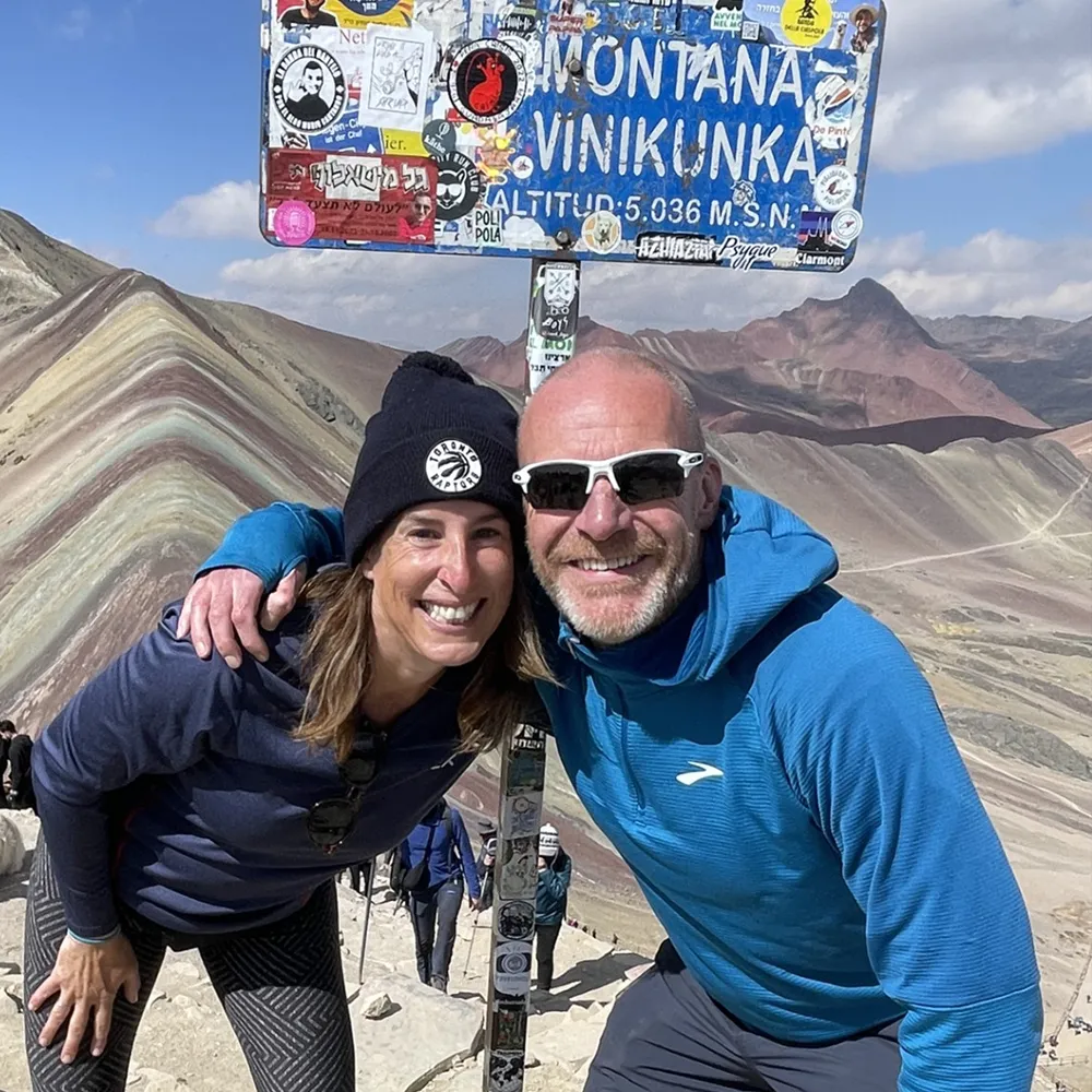 Two hikers smiling at the summit of Vinicunca, also known as Rainbow Mountain, standing beside the colourful altitude sign with the mountain’s distinctive striped landscape in the background.
