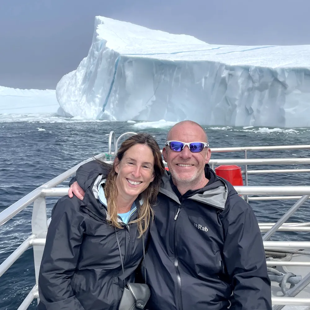 Smiling couple sitting on a boat in icy waters, with a large white iceberg towering behind them.