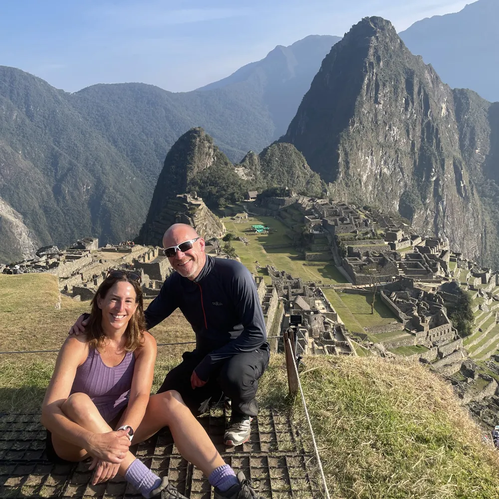 Two travellers sitting and smiling at a viewpoint overlooking Machu Picchu, with the ancient terraces and dramatic mountain peaks in the background.