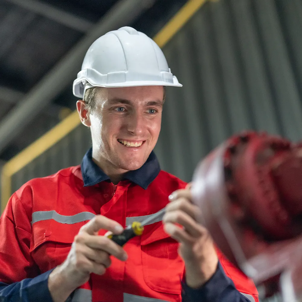 Oil and gas worker wearing a red protective uniform and white hard hat, smiling while using a tool to work on industrial machinery.