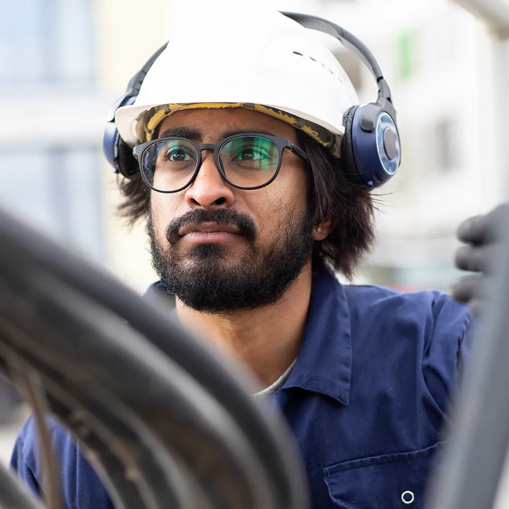 Engineer wearing a hard hat, safety ear defenders, and glasses, focusing on machinery or equipment while working on site.