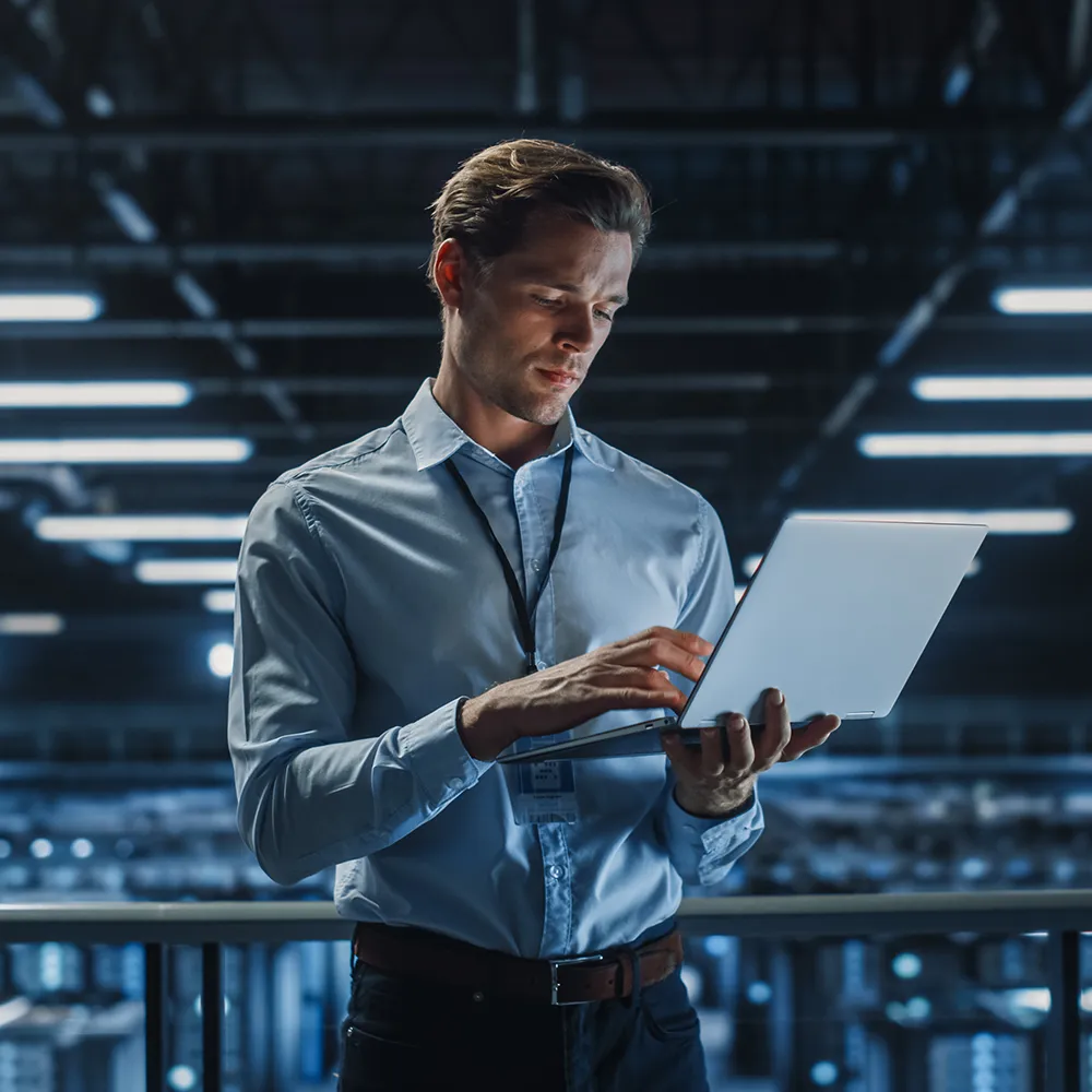 IT contractor standing in a modern data centre, using a laptop while surrounded by rows of illuminated server racks.