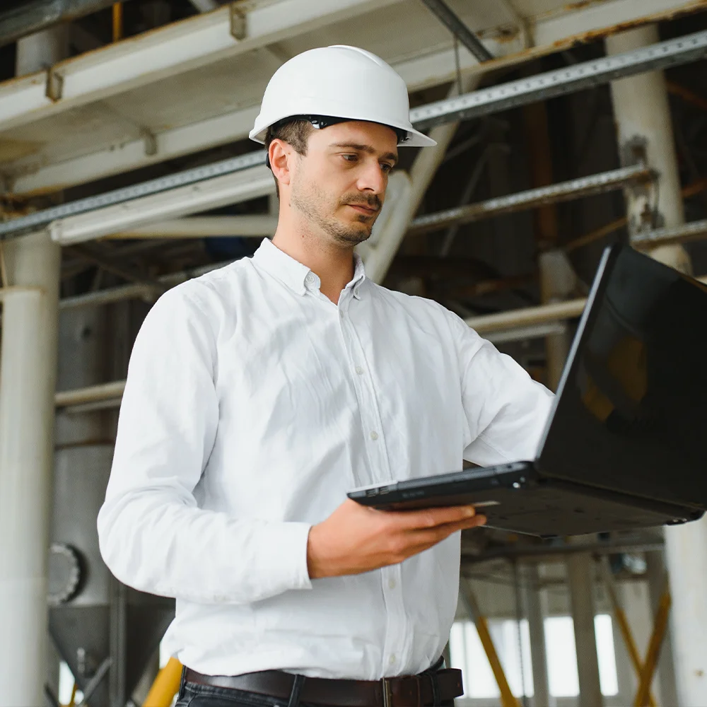 Engineer wearing a white hard hat and shirt, standing in an industrial setting while reviewing information on a laptop.