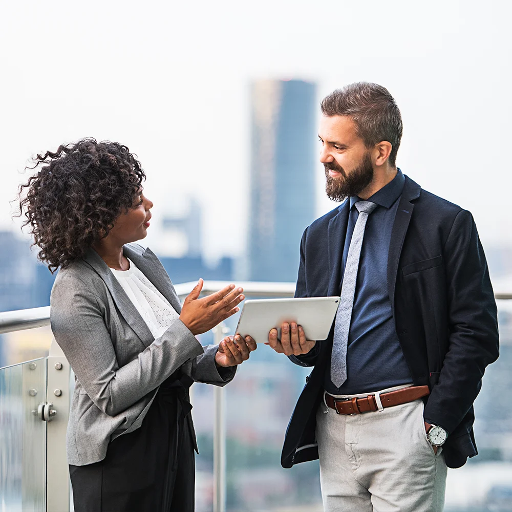 Two business professionals talking on an outdoor balcony, with one holding a tablet. They appear to be discussing work, set against a blurred city skyline in the background.