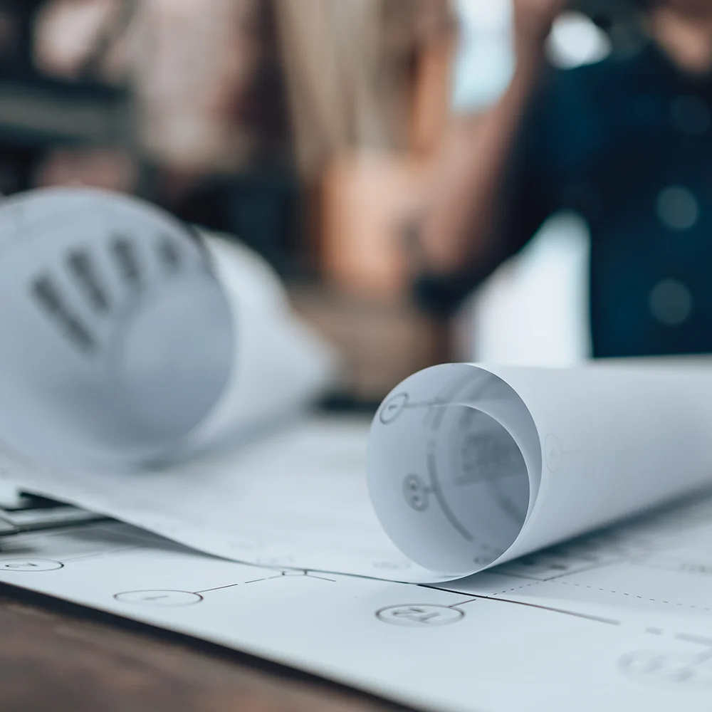 rolled-up architectural or engineering plans laid out on a desk, with a softly blurred background of people in a meeting or discussion setting.