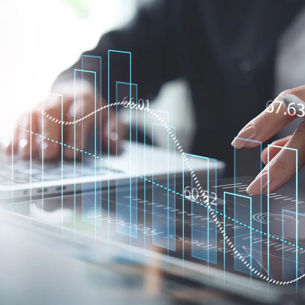 Close-up of hands working on a laptop and tablet, overlaid with transparent digital charts and graphs showing data trends and analytics.